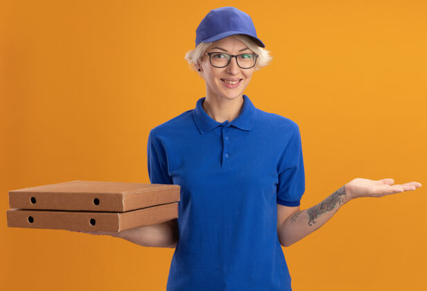 欢呼身着蓝色制服 戴着眼镜 拿着比萨饼盒的快乐的年轻送货员在橙色的墙上欢快地微笑着拿着穿着送货