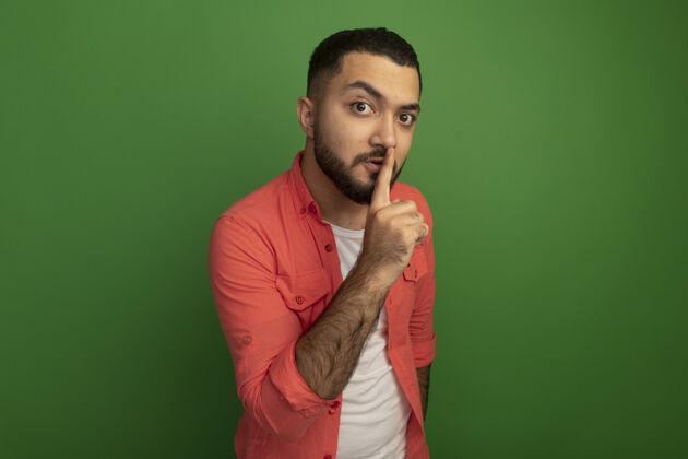 站立身穿橘色衬衫 留着胡子的年轻人站在绿色的墙上 手指放在嘴唇上做着沉默的手势沉默男人手势