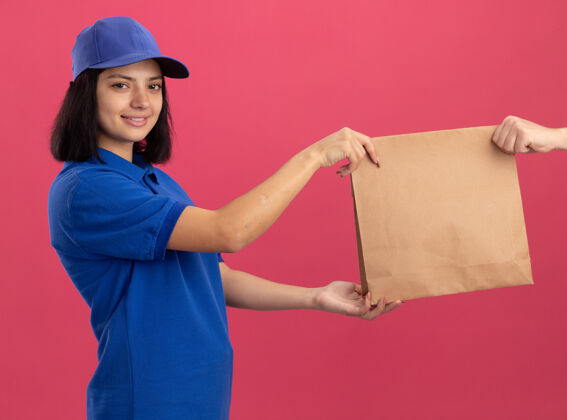 女孩身穿蓝色制服 头戴帽子的年轻送货员站在粉红色的墙上 微笑着自信地给顾客送纸包包装年轻顾客