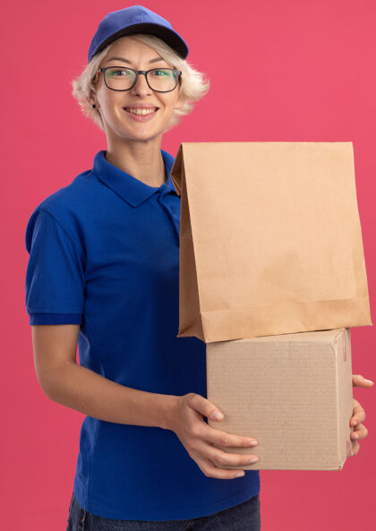 制服穿着蓝色制服 戴着帽子 手里拿着纸包和纸板箱的快乐的年轻女送货员在粉红色的墙上欢快地笑着抱着帽子送货