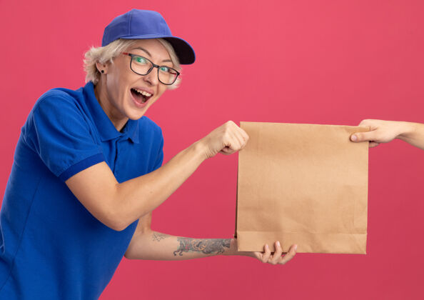 女人身穿蓝色制服 戴着帽子的年轻送货员站在粉色的墙上 一边接受纸包裹 一边兴奋不已送货帽子同时