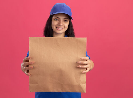 送货身穿蓝色制服 头戴鸭舌帽 拿着纸包的年轻送货女孩站在粉红色的墙上 自信地微笑着抱着制服站着