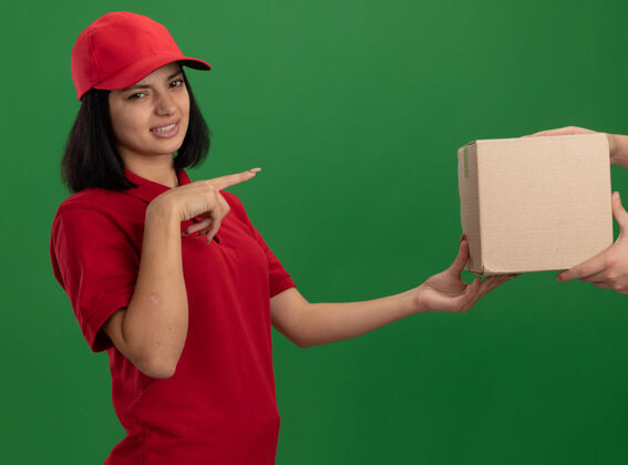 目录穿着红色制服 戴着帽子的年轻女送货员把纸板箱递给一个困惑和不高兴的顾客 用食指指着绿色墙壁上的东西女孩站手指