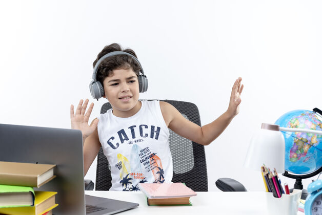 耳机高兴的小男生戴着耳机坐在书桌旁 带着学习工具 在白色背景上听孤立的音乐小男孩书桌请穿