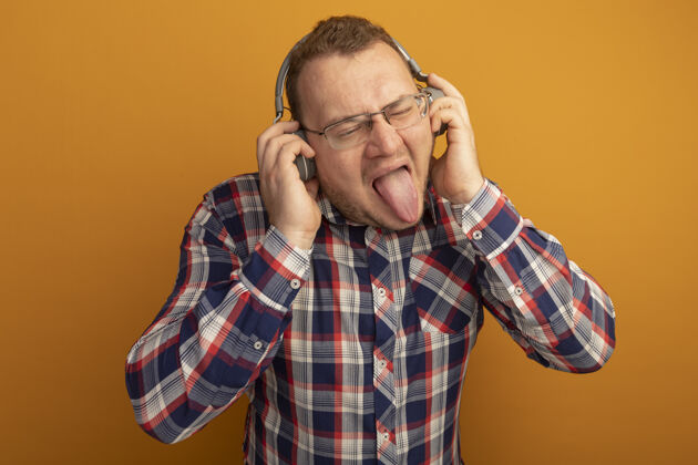 耳机戴着眼镜 穿着格子衬衫 戴着耳机的快乐男人站在橙色的墙上伸出舌头站着坚持检查