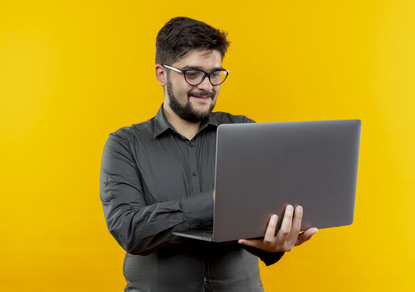 请拿着高兴的年轻商人戴着眼镜拿着和用过的笔记本电脑隔离在黄色背景上年轻人商务人士眼镜