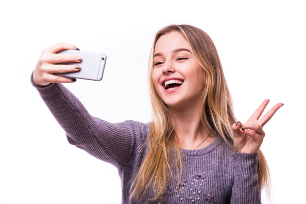 自拍一个微笑的可爱的女人在智能手机上自拍的照片被隔离在白色的墙上胜利手指肖像