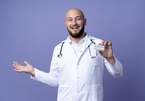 喜悦快乐的年轻秃头男医生穿着医用长袍和听诊器拿着药片 用手指着隔离在蓝色背景上的一侧穿壁板医生