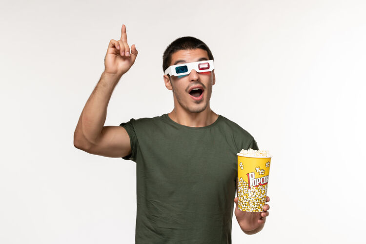 视图正面图身穿绿色t恤的年轻男子戴着d型太阳镜拿着爆米花在白墙上看电影孤独电影院男电影观看电影院电影
