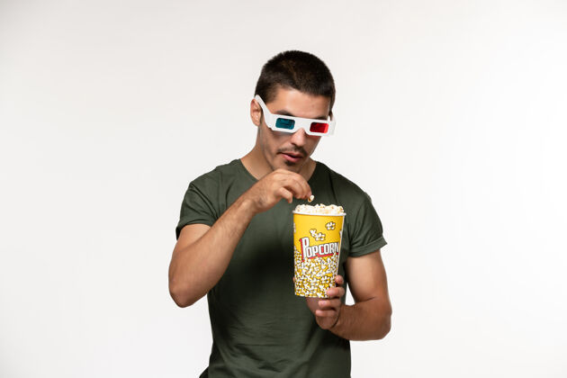 男人正面图身着绿色t恤 戴着d型太阳镜 手持爆米花的年轻男性在白色墙上拍摄电影《孤独电影院》男性电影观点肖像年轻男性