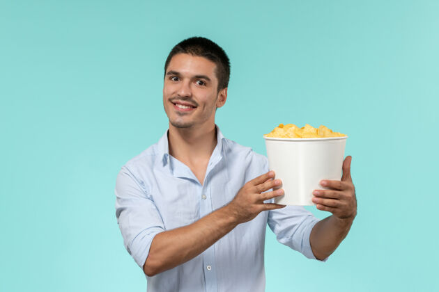 肖像正面图：年轻人拿着土豆片看电影 微笑着站在蓝色的墙上孤独地看电影看电影年轻人