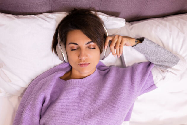 休息快乐的女人在家舒适的床上穿着暖和的衣服套头衫 听音乐年轻枕头冷静