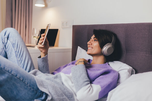 漂亮快乐的女人在家舒适的床上穿着暖和的衣服套头衫 听音乐卧室冷静科技
