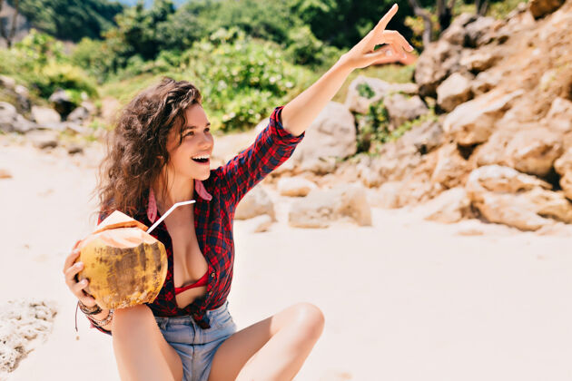 放松兴奋的快乐女人穿着短裤和明亮的衬衫坐在沙滩上 椰子鸡尾酒和指指点点阳光夏天快乐