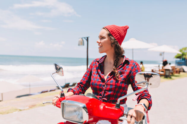 沙滩穿着格子衬衫 戴着红帽子的时髦漂亮女士在阳光下驾驶着红色的自行车 背景是蓝色的海水身体自行车阳光