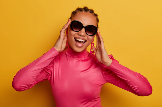 套头衫快乐的黑皮肤年轻女子的画像戴着时髦的太阳镜和粉色的毛衣 享受阳光明媚的日子 穿着鲜艳的衣服 隔离在黄色的墙上人 时尚和风格的概念外观肖像欢乐