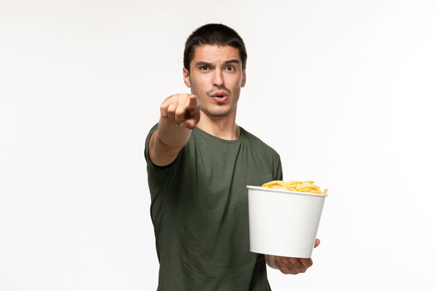 杯子正面图身穿绿色t恤的年轻男性手持土豆cips 在浅白的墙上看电影孤独的电影观看土豆电影