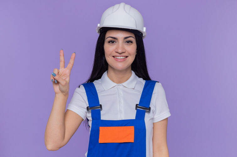 女人身穿施工制服 头戴安全帽的年轻建筑工人望着前方 兴高采烈地微笑着 紫色的墙上立着v字标志安全头盔建筑工人