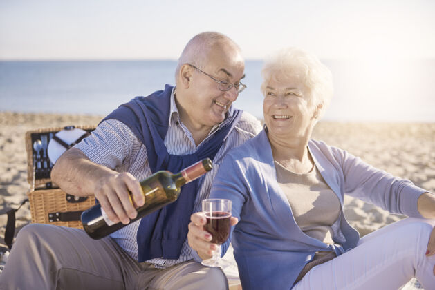 女性老人在沙滩上倒红酒 退休和暑假的概念灰色头发快乐舒适
