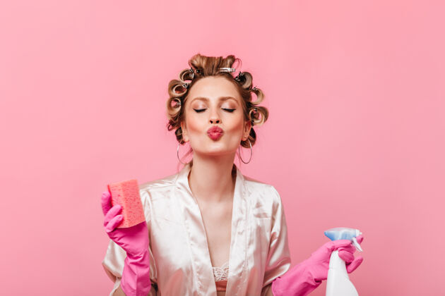 情感头上戴着卷发器的女人拿着洗碗用的海绵在粉红色的墙上吹吻肖像美丽时尚