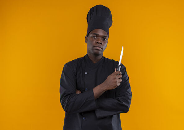 非裔美国人身着厨师制服的年轻自信的美国黑人厨师交叉双臂持刀看着橙色的相机 并留有复印空间制服交叉刀