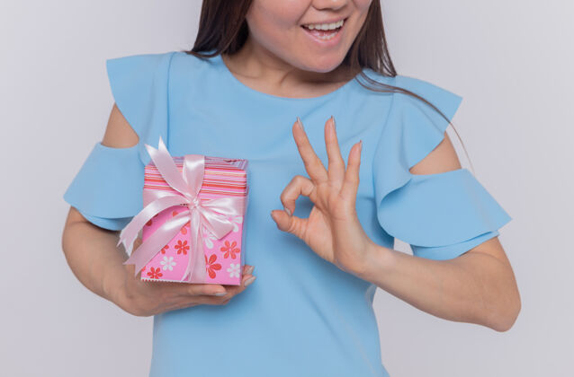 女人身着蓝色连衣裙的快乐亚洲女士手持礼物 展示ok标志 微笑着站在白墙上庆祝国际妇女节微笑快乐国际