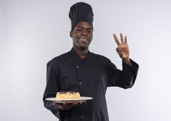 烹饪身着厨师制服 面带微笑的年轻美国黑人厨师拿着盘子上的蛋糕 在白色的空白处做手势“ok”手势标志持有厨师