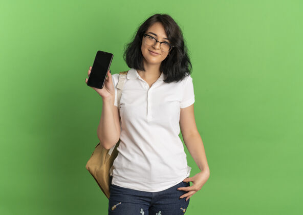 持有年轻漂亮的高加索女生戴着眼镜 背着书包拿着手机放在绿色和复印空间上空间电话请