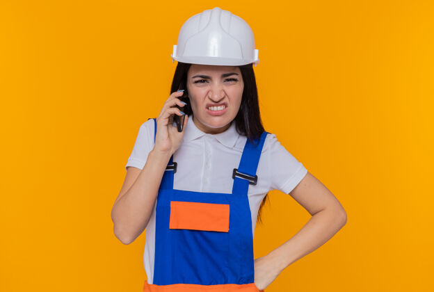 建筑工人身穿施工制服 头戴安全帽的年轻建筑工人站在橙色的墙上 一边用手机说话 一边显得既恼火又不高兴年轻人站着安全