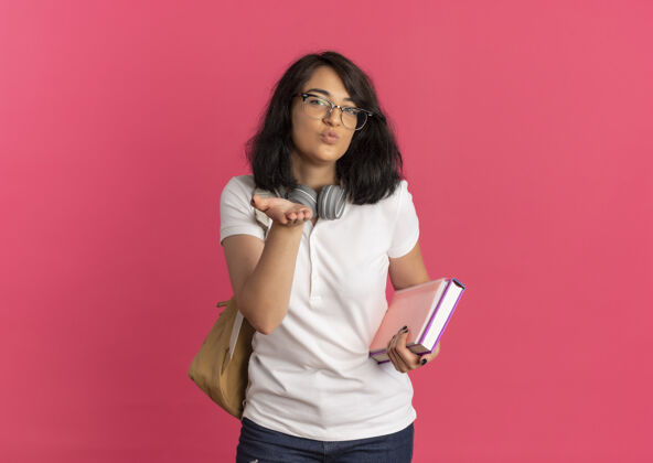空间年轻快乐漂亮的高加索女学生戴着眼镜 背着一个戴着耳机的袋子 用手拿着粉红色的书和复印空间送吻漂亮背穿