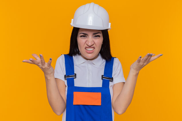 手臂身穿建筑制服 头戴安全帽的年轻建筑女工人站在橘色的墙上 看着前面恼怒地举起双臂站立恼怒建筑工人
