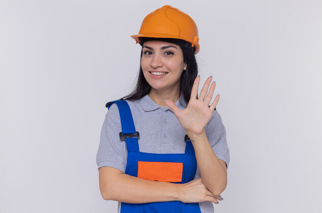 快乐身穿施工制服 头戴安全帽的年轻建筑女工人正快乐而积极地看着前面 张开的手掌站在白色的墙上开放展示手掌