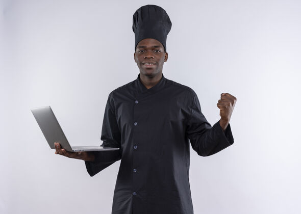 烹饪年轻的非洲裔美国厨师身着厨师制服 面带微笑 手里拿着笔记本电脑 握紧拳头对着白色 留着复印空间厨师空间年轻
