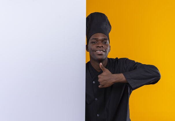 复制年轻自信的美国黑人厨师身着厨师制服 站在白色墙壁后 竖起大拇指 站在橙色的空间里橙色立场非裔美国人