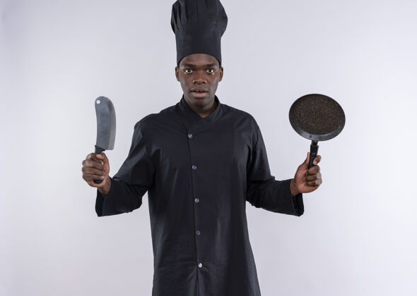 烹饪穿着厨师制服的年轻的非洲裔美国厨师拿着刀和煎锅 在白色的地板上留有复印空间制服复制年轻