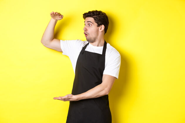 厨师穿着黑色围裙的侍者看着一个大东西 手里拿着一个大东西 站在黄色的背景上工作室站立尺码