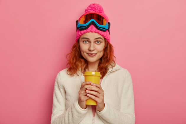 帽子迷人的红发女人戴着暖和的帽子 滑雪镜和白色套头衫满意茶一次性