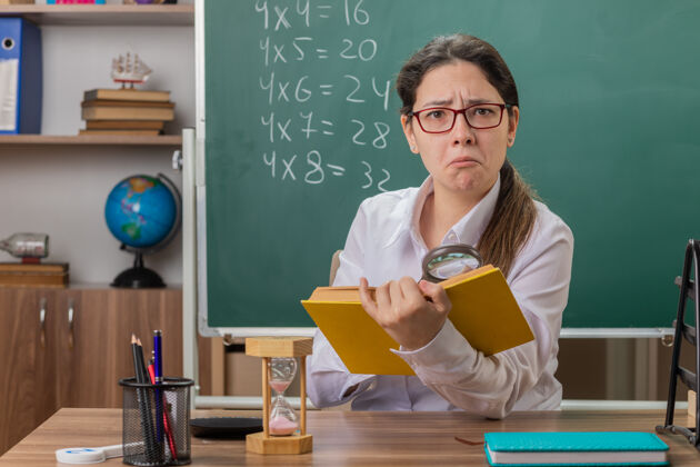 女人年轻的女老师戴着眼镜拿着书看着放大的书页坐在教室黑板前的课桌上困惑不已拿着书桌坐着