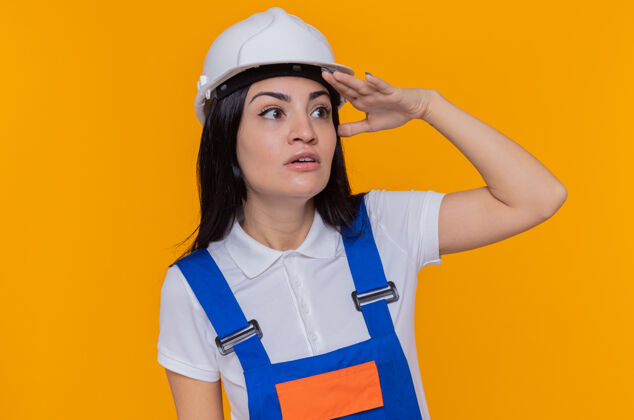 很远身穿施工制服 头戴安全帽的年轻建筑女工人远远望着远处 用手捂着头 看着橘色的墙上站着什么人或什么东西手安全站着