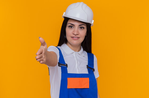 安全身着施工制服 头戴安全帽的年轻建筑工人站在橙色的墙上 微笑着自信地向前方致意站立微笑自信