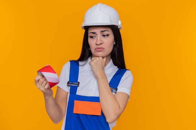 手身穿建筑制服 戴着安全帽 手放在下巴上看着胶带的年轻建筑工人站在橙色的墙上思考头盔建筑工人制服