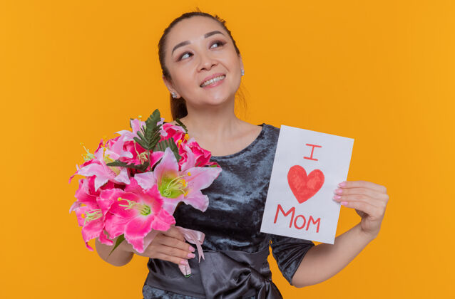站立快乐快乐的亚洲女性母亲手持贺卡和花束庆祝母亲节 站在橙色的墙上 抬头微笑着快乐举办花