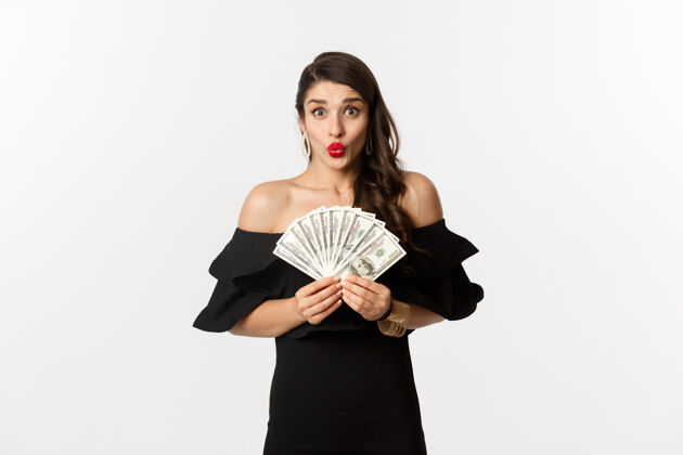 优雅时尚和购物概念兴奋的女人穿着黑色连衣裙 红唇 展示金钱美元 看着相机 白色背景美丽魅力服饰