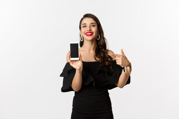 优雅网上购物概念穿着黑色连衣裙的时尚女性用手指着智能手机屏幕 展示应用程序 站在白色背景上女性美丽女性