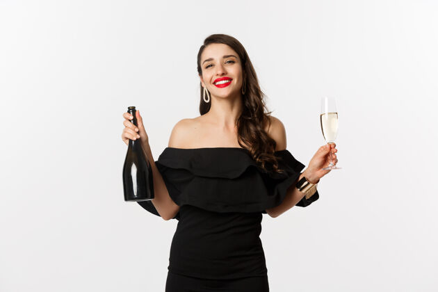 女性庆典和派对概念穿着迷人礼服的时尚黑发女子 手持一瓶香槟和一杯香槟 高兴地微笑着 站在白色背景上黑发美丽女人