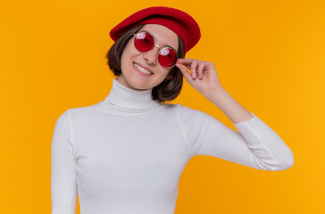 年轻快乐而积极的年轻女子 短发 白色高领毛衣 头戴贝雷帽 戴着红色太阳镜 站在橘色的墙上 面带微笑地看着前方微笑积极快乐