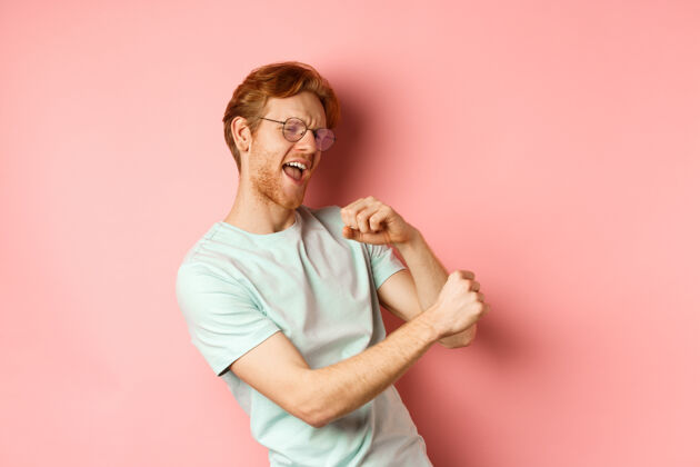 人暑假的概念快乐的年轻人 红头发 跳舞和娱乐 唱歌的音乐 站在粉红色的背景眼镜表情男人