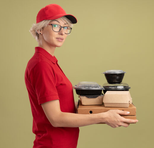 帽子身穿红色制服 戴着眼镜 手持比萨饼盒和食品包的年轻送货员站在绿色的墙上 自信满满制服穿着站着
