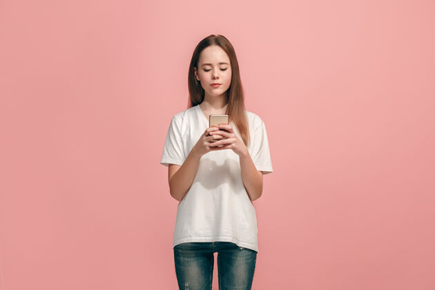女人快乐的少女站着 微笑着用手机在时尚的粉色工作室背景上美丽的女性半身像站电话表情