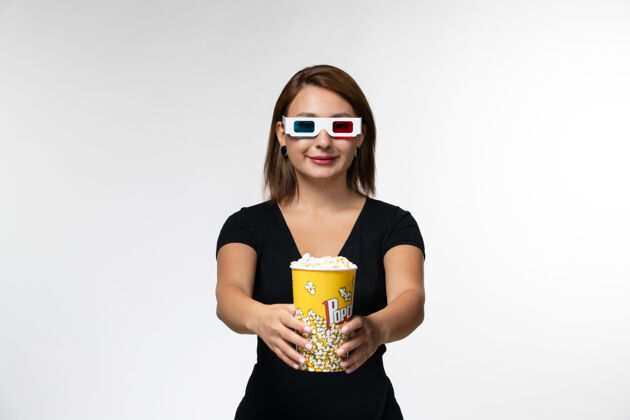 漂亮正面图年轻女性戴着d型太阳镜拿着爆米花看电影 在白色表面微笑爆米花前面电影